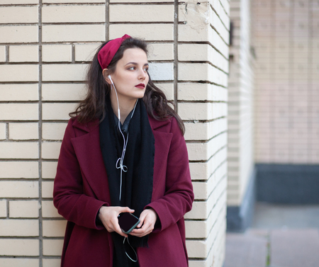 young brunette woman in coat listen music with wire headphones near brick wall on autumn streetの写真素材