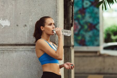 young woman in sportswear drinking water near concrete wall after outdoor workout in city