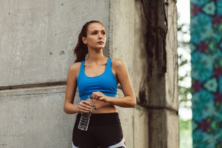 young woman in sportswear with bottle of water standing near concrete wall, outdoor workoutの写真素材