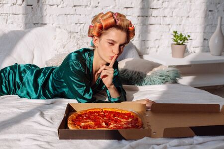 young woman in silk pajama showing silence gesture with pizza pepperoni in bedの写真素材