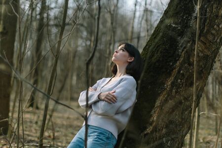 young thoughtful woman standing near tree in wild forest, looking awayの写真素材