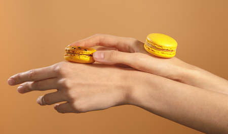 two tasty yellow dessert macarons on female hands isolated on brown backgroundの写真素材