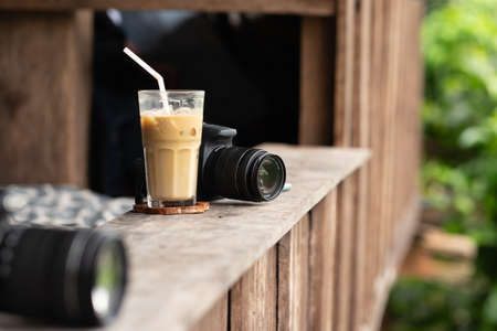Black camera behind a glass of iced coffee put on small wood tabletop at balcony edge. Focus on camera. Coffee, cafe or travel concept.の写真素材