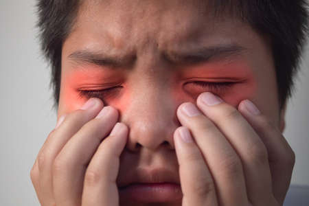 The boy pains his eyes. He massages his eye by fingers. This shot is close up portrait. At red color, it shows position of symptoms.の写真素材