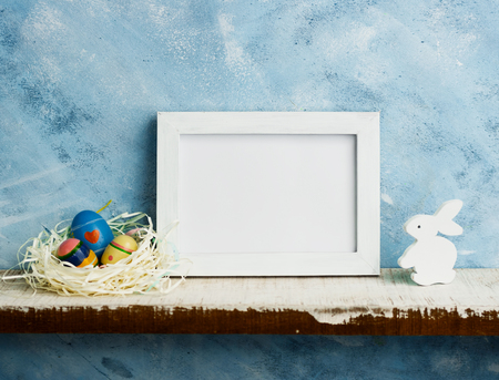 Eatser mock up with photoframe, painted eggs in nest and white wooden bunny on shelf against blue wall. Scandinavian style. Text spaceの写真素材