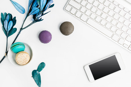 Top view flat lay colorful macarons with keyboard, cell phone and blue leaves on white table. Creative dessert concept, text spaceの写真素材