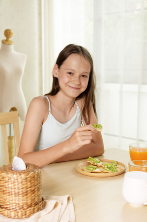 Cute smiling preteen girl having healthy breakfast: avocado sandwich and orange juice. Healthy lifestyle concept, vegetarian food. Superfoods. Living room interior in backgroundの写真素材