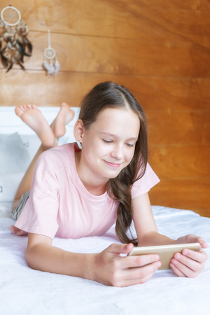Cute preteen girl in pink t-shirt and shorts lying on bed with cell phone in boho style room against wooden wall and dreamcatchers. Scandinavian interior, modern lifestyle concept. Text spaceの写真素材