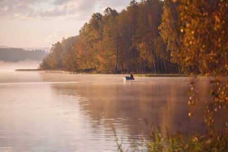 Fisherman in a boat on autumn lake in morning lightの写真素材