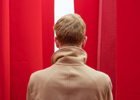 Man in a coat peers out from behind red curtainsの写真素材