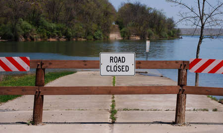 Warning sign and barrier where old highway disappears into a lake and reappears from the water in the background.の写真素材