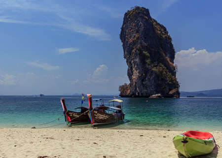 Krabi, Thailand- March 29 2019:traditional wood long tail boat in tirquese blue water, and green kayak in white beach sand, islands tour, huge rock backgroundのeditorial素材
