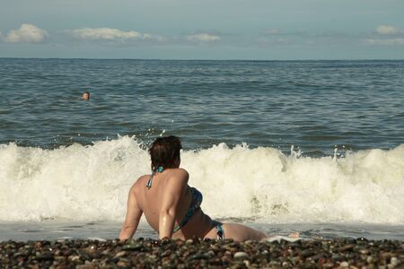 Back view of adult woman in colorful swimsuit that sitting on shore of the sea on the beach see a wavesの写真素材