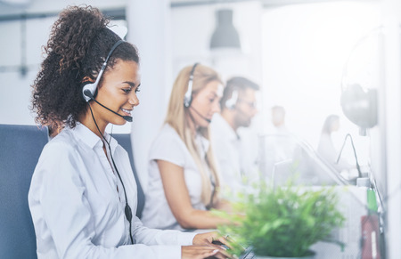 Call center worker accompanied by her team. Smiling customer support operator at work. Young employee working with a headset.の写真素材