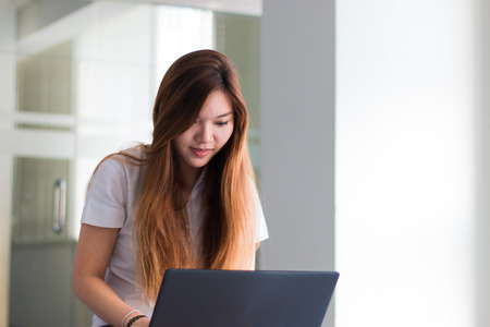 Asian woman student working on a laptop smiling in building near window with sunlightの写真素材