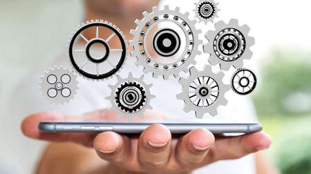 Businessman on blurred background holding floating gear icons over mobile phone 3D renderingの写真素材