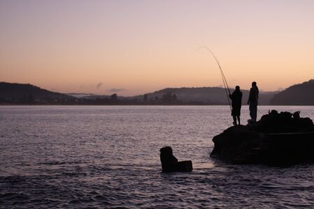 Dawn silhouette of fishermen on rocksの写真素材