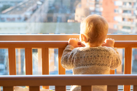 A child looks at the city through a large window while standing in a cribの写真素材