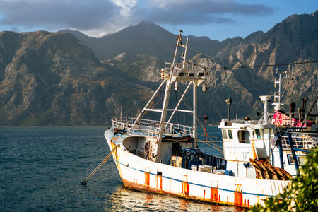 An old fishing boat rests by the calm sea with towering mountains behind, showcasing a serene and timeless coastal scene.の写真素材