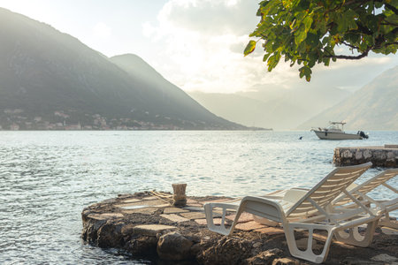 A tranquil seaside spot with lounge chairs on a stone pier, overlooking calm waters, majestic mountains, and a small boat in the distance.の写真素材