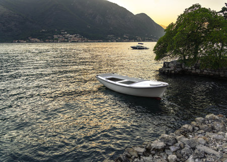 A small white boat gently floating on calm waters by a rocky shore, framed by trees and mountains under the glow of sunset.の写真素材