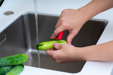 Peeling a fresh cucumber under running water with a red peeler, highlighting healthy and clean food preparation in the kitchen.の写真素材