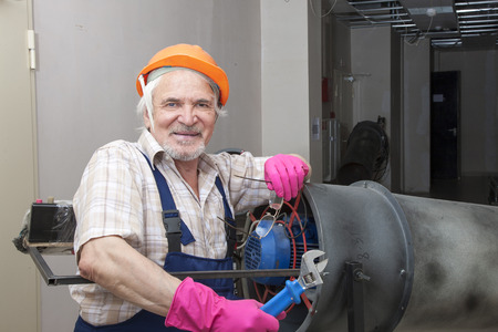 Elder man fixing an industrial heater in orange helmet and pink gloves with wrenchの写真素材