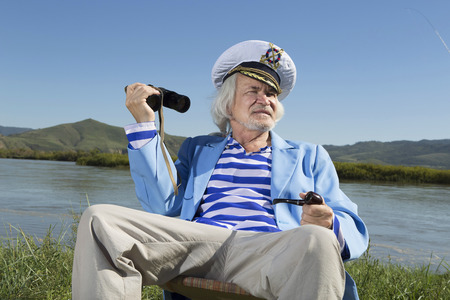 The captain on vacation looking through binoculars while sitting on the river bankの写真素材