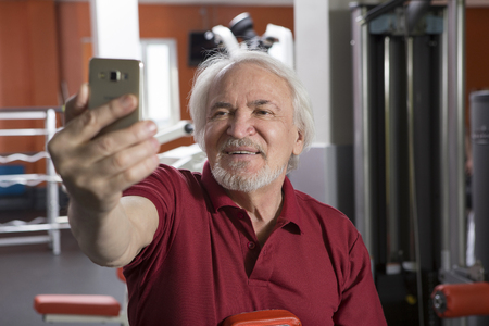 Elderly man taking a selfie with smartphone in fitness centerの写真素材