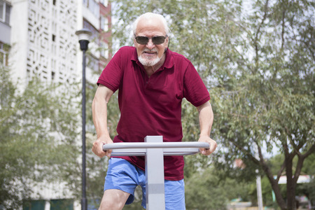Attractive senior man trains on sporting equipment in a city in the open air. The concept of a healthy lifestyle and accessibility of sports training for every person. Available sports equipment in a public place.の写真素材