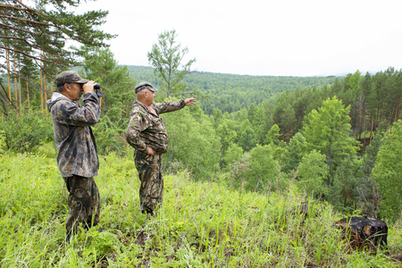 Foresters went to the protection of the forest. An adult male forester examines the forest from the top of the mountain through binoculars. The huntsman shows the directions of the brown bear habitat.の写真素材