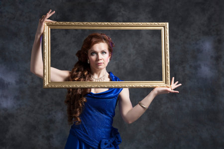 Young woman in blue classic evening dress posing in the studio on a gray background with a frame for...