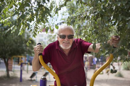 Attractive senior man trains on sporting equipment in a city in the open air. The concept of a healthy lifestyle and accessibility of sports training for every person. Available sports equipment in a public place.の写真素材