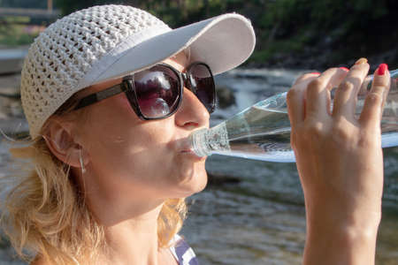 Yaremche Ukraine. 25 June 2021: Young beautiful girl with closed eyes in a baseball cap drinks water on the background of the river.のeditorial素材