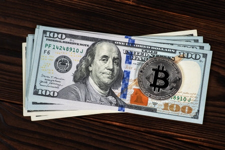 Silver symbolic coin Bitcoin on stack banknotes of one hundred dollars on wooden background. Exchange bitcoin cash for a dollars. Cryptocurrency on US dollar bills. Digital modern method of payment.の写真素材