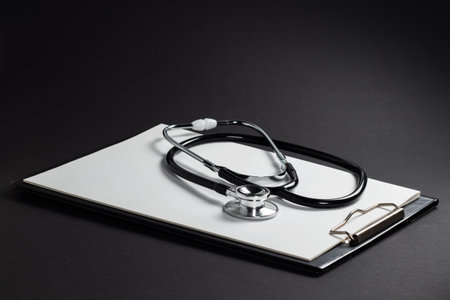A clipboard and stethoscope with white paper on it, set against a dark background.の写真素材