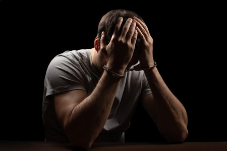 Man, criminal and handcuffs for interrogation, crime or money laundering in arrest on table in room. Closeup of male person, gangster or suspect in jail, justice or guilty for fraud, theft or scamの写真素材