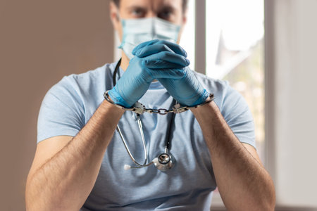 A medical officer, doctor, or quack in a blue medical uniform in handcuffs. The concept of crimes committed by medical personnel or providing medical services without a license.の写真素材