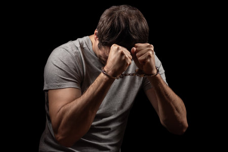 Handcuffed man covering his face with his hands. Arrested and handcuffed criminal man with handcuffs on his hands cover his head and regret in the police station.の写真素材