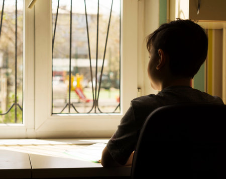The quarantined boy sadly looks at the street through the window, there is COVID-19