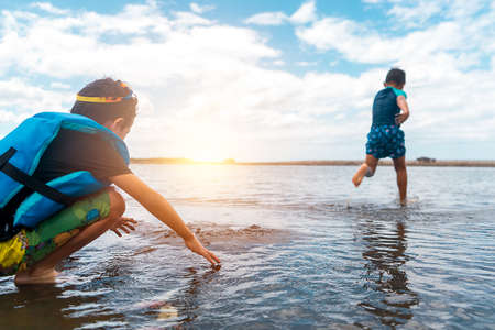 Two children playing on the beach at sunset. One of them has a life jacket and the other runs to dive into the seaの写真素材