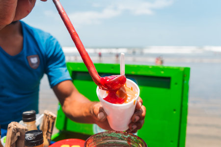 Close-up of a street vendors hand spilling gooseberry candy onto shaved ice at the beach.