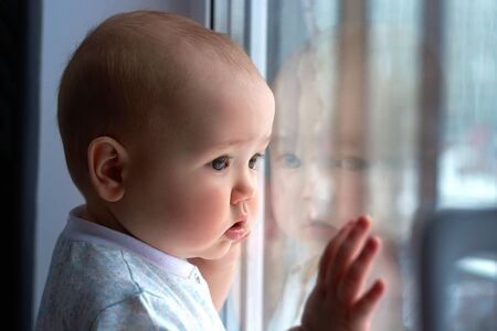 a small boy on the windowsill looks out the window.a child with a sad expression on his faceの写真素材