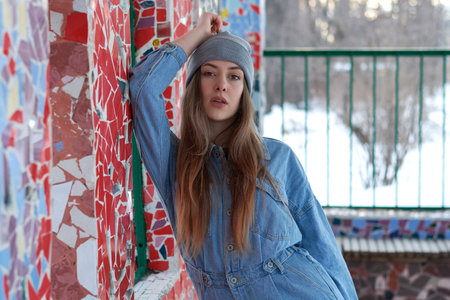a woman in blue denim overalls and a gray hat stands against a wall of colored glass.