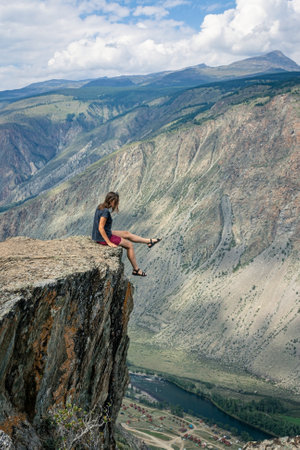 Dangerous photos in nature. A woman sits on the edge of a sheer cliff above a high cliff and dangles her legs. fatal risk for photography. Summer Altai, Chulyshman valleyの写真素材