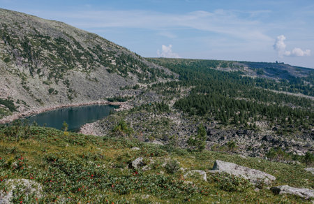 Hills strewn with large stones, a blue lake below, Coniferous trees in green forest and rocks on the horizon. Summer landscape of wild nature in low mountains.の写真素材