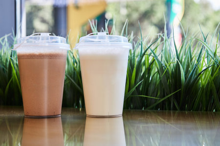 Two clear plastic glasses with lids filled with chocolate vanilla milkshakes on table, green grass. Blank mockup, background beverage cafe. Take away milkshake, coffee shop at gas station. Copy spaceの写真素材