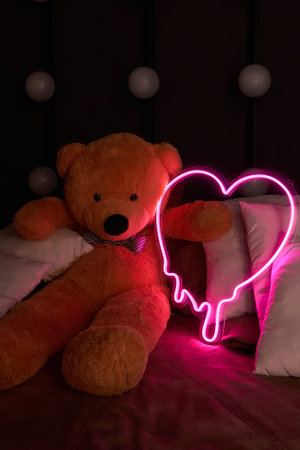 Brown teddy bear lies in pillows and holds a neon pink heart. Valentine's Day 14 February, Gift romantic background. Declaration of love, congratulations on the holiday or anniversary.の写真素材