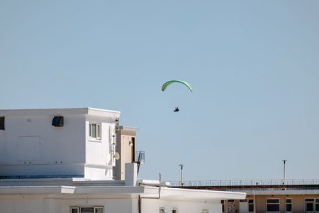 Paraglider mid-flight against clear blue sky in city. Paraglider's canopy is a vibrant green and...