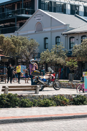 Man standing next to motorcycle and wearing a helmet in front of an old white building at Hermanus Waterfront complex in South Africa. Urban travel and tourism. Cape Town, South Africa - 25 OCT 2023のeditorial素材